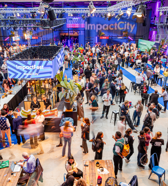 A vibrant indoor event at ImpactFest, hosted by ImpactCity, with a large crowd of people networking, eating, and engaging at various booths and tables. The space is well-lit with purple and blue lighting, featuring banners, food stalls, and seating areas. Overhead trusses hold lights and signs, and the venue has a modern industrial design with high ceilings and visible catwalks.
