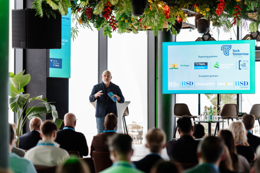 A speaker on stage at the Tech Tomorrow event with an audience and a presentation on the screen.