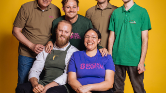 A group of six people posing together in front of a yellow background.