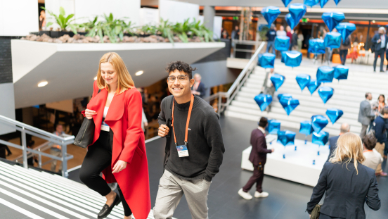 A smiling young man and a woman in a red coat walk up a staircase at a busy indoor event, with floating blue diamond-shaped balloons in the background.