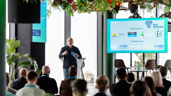 A speaker on stage at the Tech Tomorrow event with an audience and a presentation on the screen.