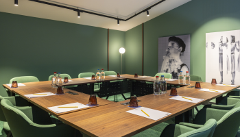 A meeting room with a wooden table, green chairs, water bottles, and notepads. Two artworks on the wall.