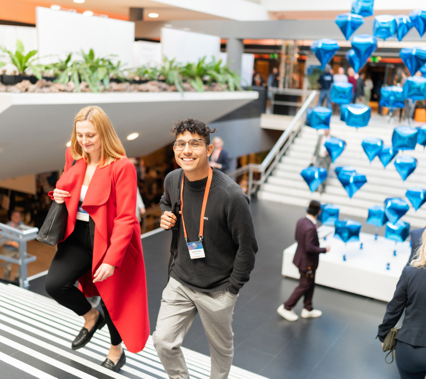 A smiling young man and a woman in a red coat walk up a staircase at a busy indoor event, with floating blue diamond-shaped balloons in the background.