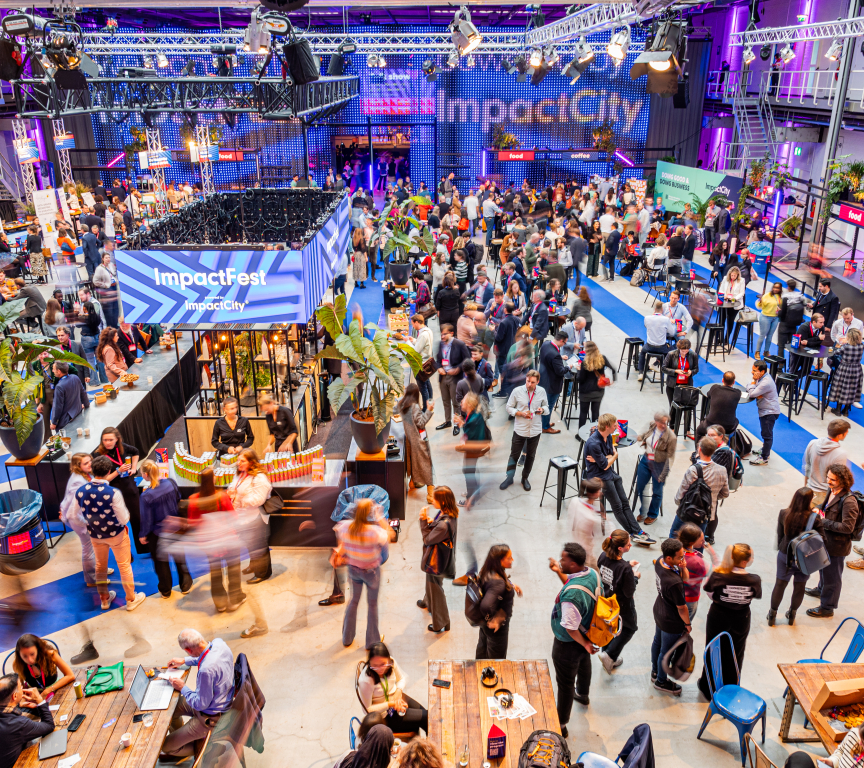 A vibrant indoor event at ImpactFest, hosted by ImpactCity, with a large crowd of people networking, eating, and engaging at various booths and tables. The space is well-lit with purple and blue lighting, featuring banners, food stalls, and seating areas. Overhead trusses hold lights and signs, and the venue has a modern industrial design with high ceilings and visible catwalks.