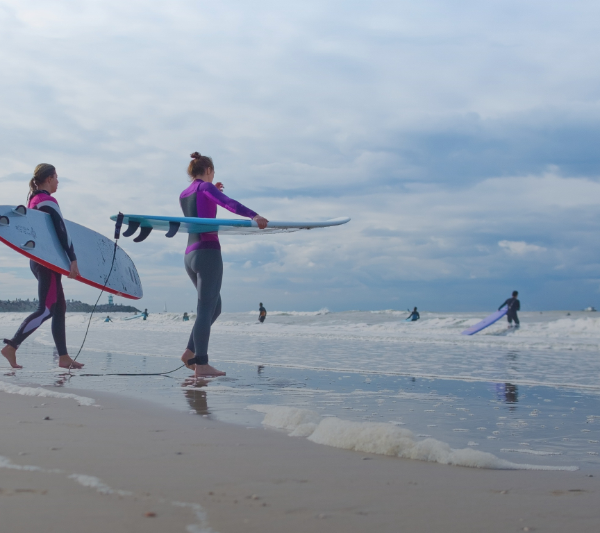 Two surfers with boards walk along the water.