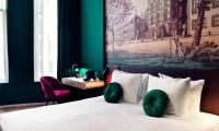 A stylish hotel room featuring a bed with green cushions, a lit bedside table, and a wall artwork.