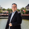 A smiling man stands by the water with flags in the background.