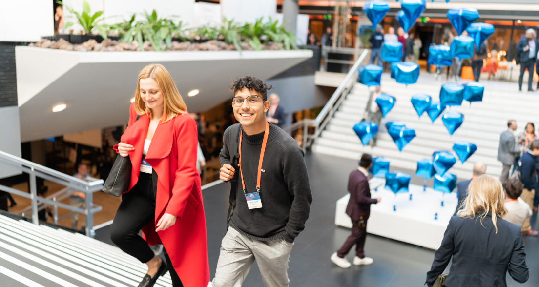 A smiling young man and a woman in a red coat walk up a staircase at a busy indoor event, with floating blue diamond-shaped balloons in the background.