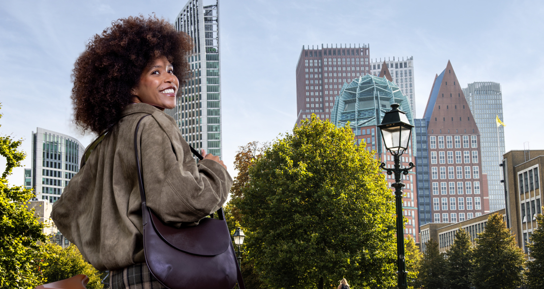 A woman with curly hair turns around and poses with a bag in a park in The Hague, with modern buildings in the background.