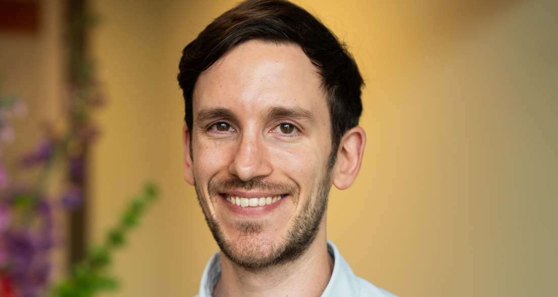 A man in a light shirt smiling, standing in front of a floral background