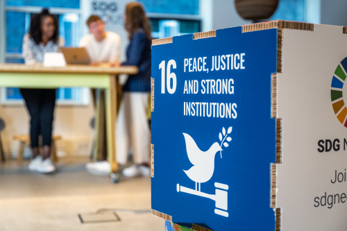 Close-up of a blue cube displaying Sustainable Development Goal 16: 'Peace, Justice and Strong Institutions,' featuring a white dove holding an olive branch perched on a gavel. In the blurred background, three people are having a discussion around a high table, suggesting a collaborative and professional setting.