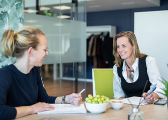 Two women in a modern meeting room, smiling and taking notes, with fruit on the table.