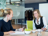 Two women in a modern meeting room, smiling and taking notes, with fruit on the table.