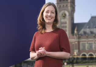 A woman in a chestnut brown sweater stands in a park with the Binnenhof in the background.