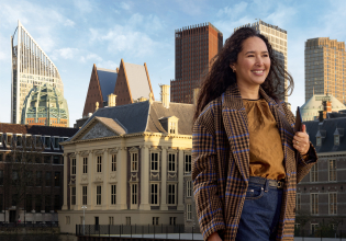 A woman in a brown coat poses by the Hofvijver, with modern buildings in the background.