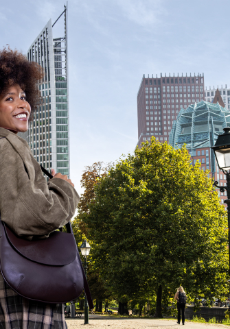A woman with curly hair turns around and poses with a bag in a park in The Hague, with modern buildings in the background.