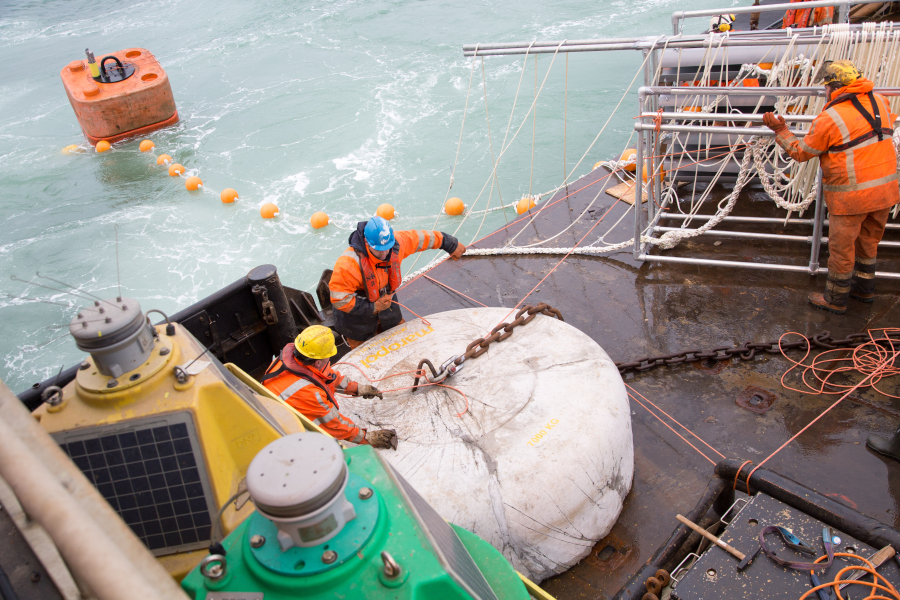 Workers in safety gear on a marine vessel handling heavy equipment and ropes as part of an offshore operation. A large concrete anchor marked '7000 KG' is being secured with chains and cables. Nearby, floating buoys and safety lines are visible in the water, indicating a marine construction or cable-laying project.