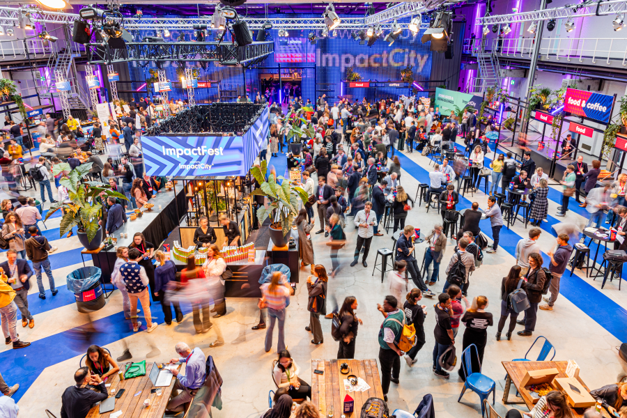 A vibrant indoor event at ImpactFest, hosted by ImpactCity, with a large crowd of people networking, eating, and engaging at various booths and tables. The space is well-lit with purple and blue lighting, featuring banners, food stalls, and seating areas. Overhead trusses hold lights and signs, and the venue has a modern industrial design with high ceilings and visible catwalks.