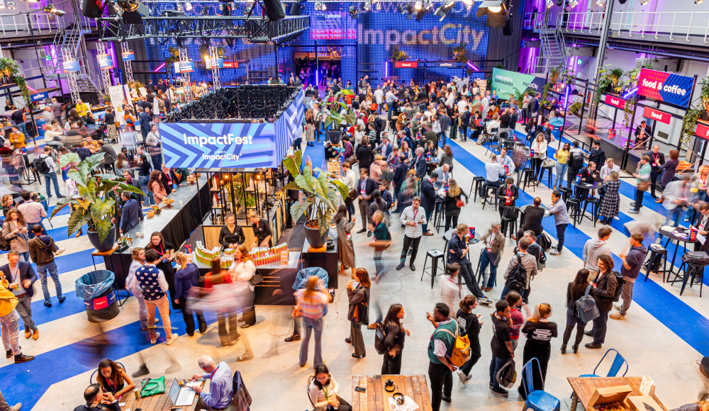 A vibrant indoor event at ImpactFest, hosted by ImpactCity, with a large crowd of people networking, eating, and engaging at various booths and tables. The space is well-lit with purple and blue lighting, featuring banners, food stalls, and seating areas. Overhead trusses hold lights and signs, and the venue has a modern industrial design with high ceilings and visible catwalks.