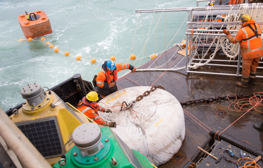 Workers in safety gear on a marine vessel handling heavy equipment and ropes as part of an offshore operation. A large concrete anchor marked '7000 KG' is being secured with chains and cables. Nearby, floating buoys and safety lines are visible in the water, indicating a marine construction or cable-laying project.