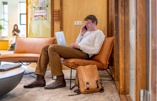 A guy in a white short working on his laptop on the couch while on the phone