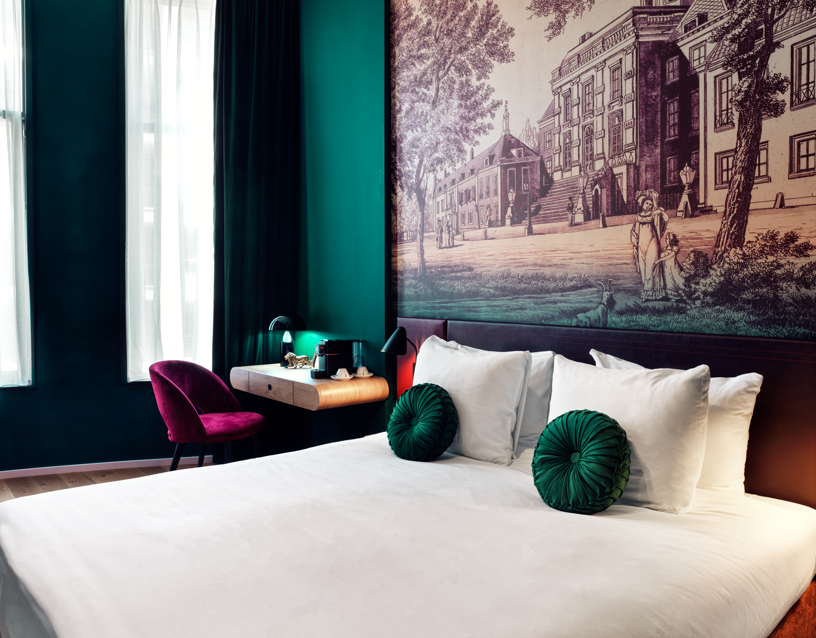 A stylish hotel room featuring a bed with green cushions, a lit bedside table, and a wall artwork.