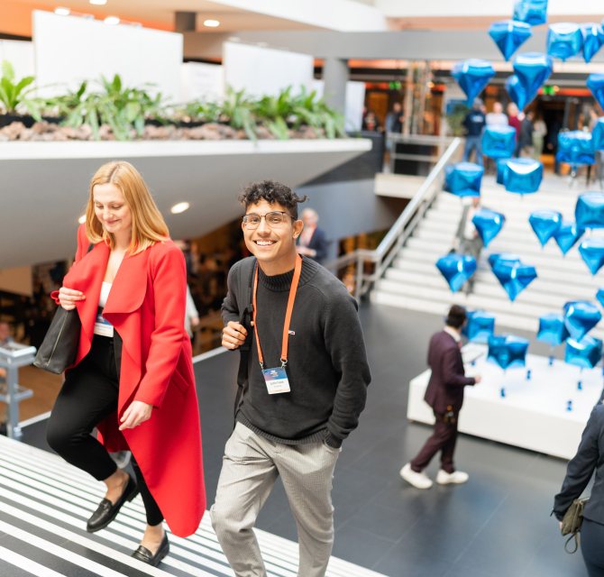 A smiling young man and a woman in a red coat walk up a staircase at a busy indoor event, with floating blue diamond-shaped balloons in the background.