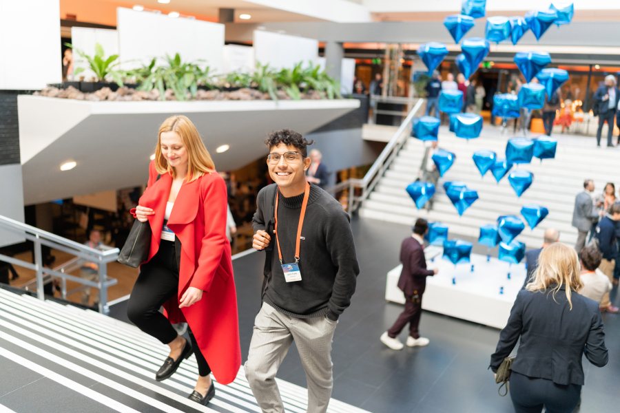 A smiling young man and a woman in a red coat walk up a staircase at a busy indoor event, with floating blue diamond-shaped balloons in the background.
