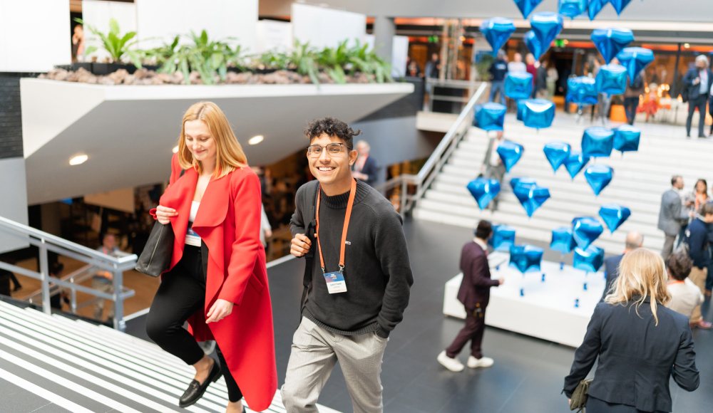 A smiling young man and a woman in a red coat walk up a staircase at a busy indoor event, with floating blue diamond-shaped balloons in the background.