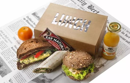 A lunchbox with sandwiches, an orange, an energy bar, and juice.