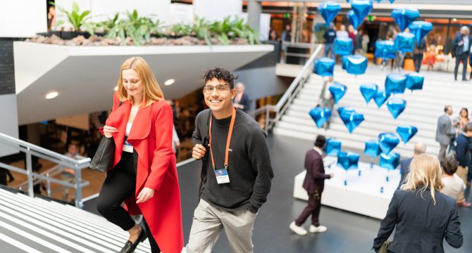 A smiling young man and a woman in a red coat walk up a staircase at a busy indoor event, with floating blue diamond-shaped balloons in the background.