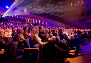 A packed theater audience watching the stage.
