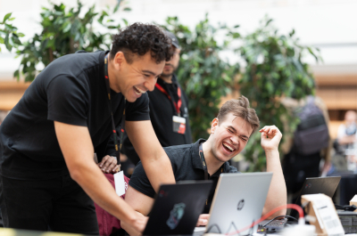 Two men are laughing and working on laptops in a green setting.