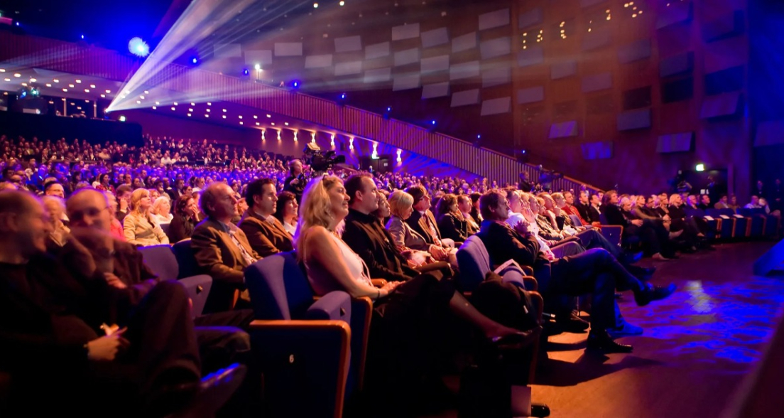 A packed theater audience watching the stage.