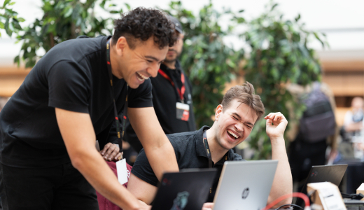 Two men are laughing and working on laptops in a green setting.