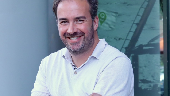 Een man met een lichtgrijze polo staat met gekruiste armen en glimlacht.
