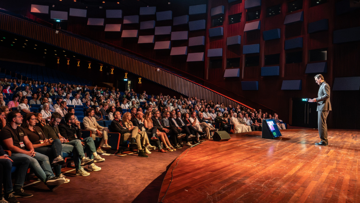 Een spreker staat op een podium voor een publiek.