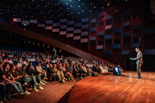 Een spreker staat op een podium voor een publiek.