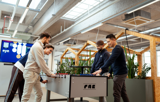 Drie mensen spelen tafelvoetbal in een moderne ruimte.
