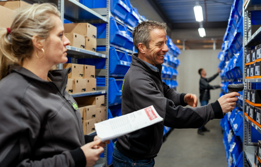 Twee werknemers in een magazijn met blauwe schappen.