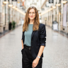 A woman in a blazer stands smiling in a shopping arcade.