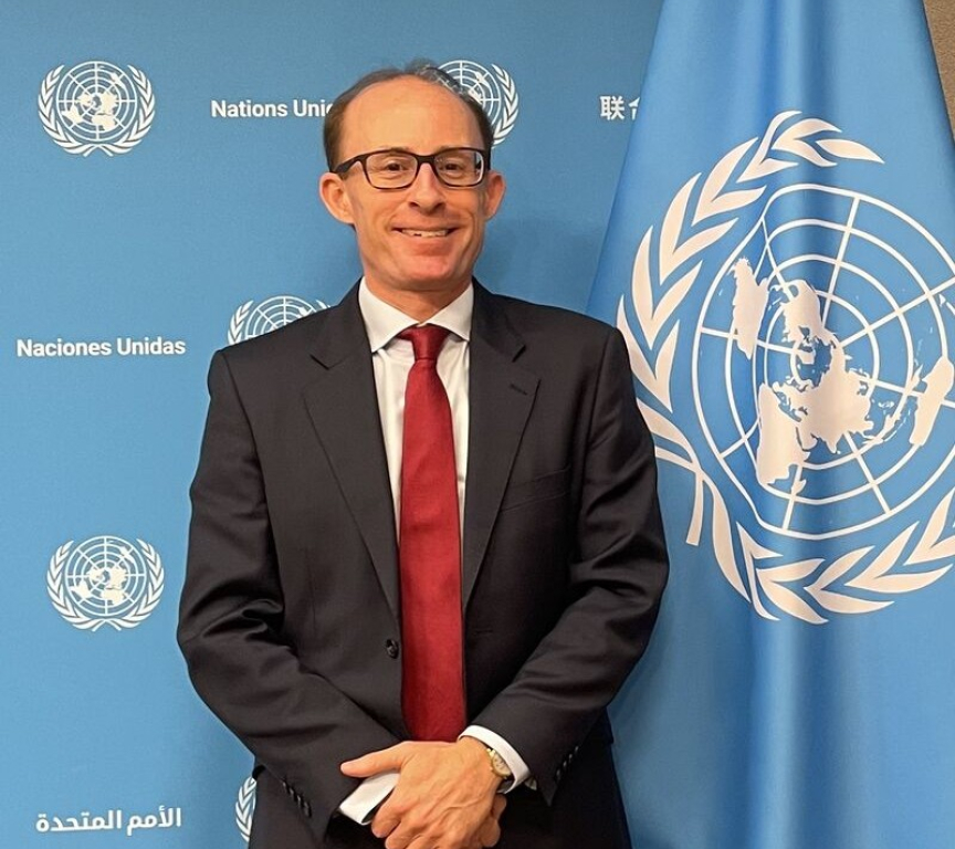 A man in a black suit with a red tie stands in front of a UN flag.