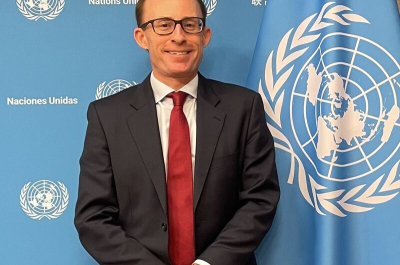 A man in a black suit with a red tie stands in front of a UN flag.