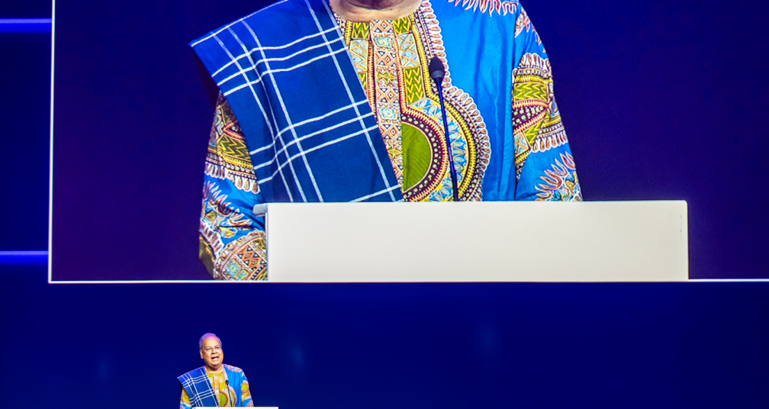 A man in traditional African attire speaks from a podium.