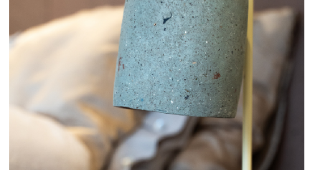 A modern lamp with a concrete shade and yellow base, on a table with cushions in the background.