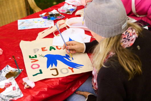 A person is painting a tote bag with the words 'Peace', 'Love', and 'Joy' and a blue bird