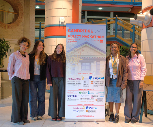 Group of young women posing next to a banner for the Cambridge Policy Hackathon.