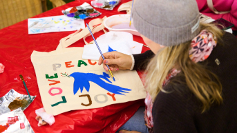 A person is painting a tote bag with the words 'Peace', 'Love', and 'Joy' and a blue bird