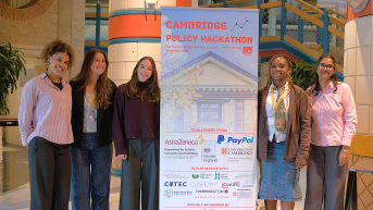 Group of young women posing next to a banner for the Cambridge Policy Hackathon.