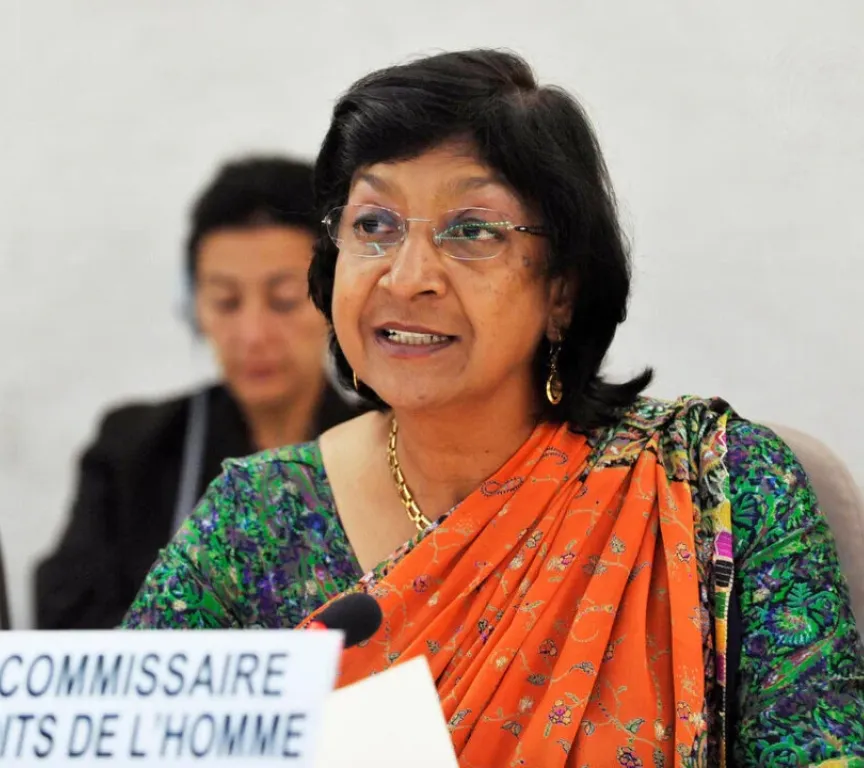 A woman in an orange and green sari speaks at a meeting.